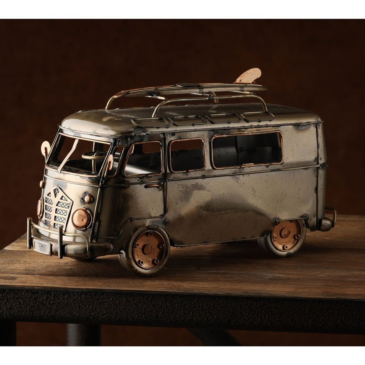 Weathered Campervan Metal Figurine with Surfboard in bronze metallic finish, featuring mesh grille, round headlamps, copper-edged windows and roof rack, shown on a wooden surface.