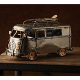 Weathered Campervan Metal Figurine with Surfboard in bronze metallic finish, featuring mesh grille, round headlamps, copper-edged windows and roof rack, shown on a wooden surface.