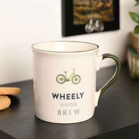 White porcelain Wheely Good Brew mug with green bicycle illustration, green rim and handle, shown on a dark surface in a home setting.