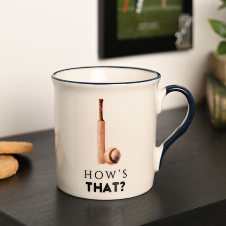 Cream porcelain mug with navy rim and handle, printed cricket bat and ball design above "HOW'S THAT?" text, shown on a dark shelf beside biscuits.