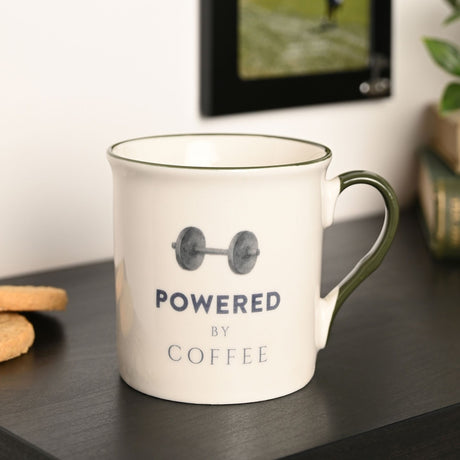 Cream porcelain mug with green rim and handle, printed with a dumbbell graphic and "Powered By Coffee" text, shown on a dark surface with biscuits nearby.