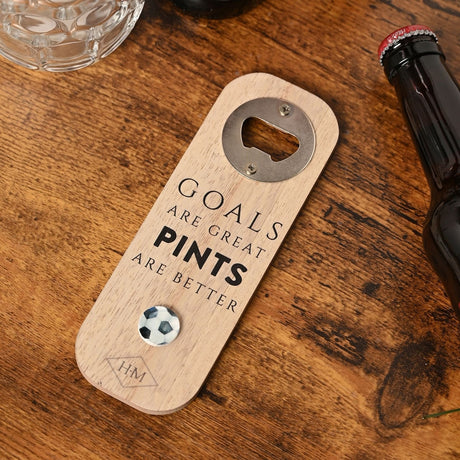 Natural wood rectangular bottle opener with metal cap-lifter plate, magnetic football disc, and printed slogan "Goals are great, pints are better," shown on a wooden table beside a beer bottle.