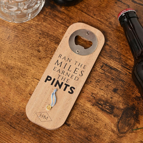 Wooden bottle opener with "Ran The Miles Earned The Pints" text and a raised blue and gold medal motif, shown on a dark wood table beside a beer bottle and glass mug.