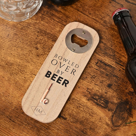 Wooden bottle opener with rounded ends in natural wood grain, metal opener screwed at the top, "BOWLED OVER BY BEER" text and cricket bat and ball motif, shown on a wooden table beside a beer bottle.