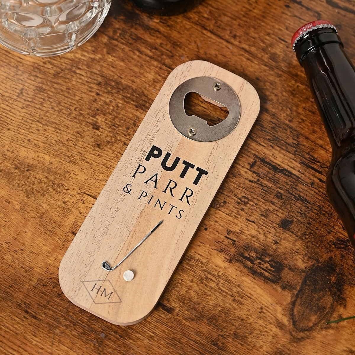 Wooden rectangular bottle opener in natural finish with "PUTT PARR & PINTS" text, a silver-toned opener plate, and a small golf club and ball detail, shown on a wooden table beside a beer bottle.