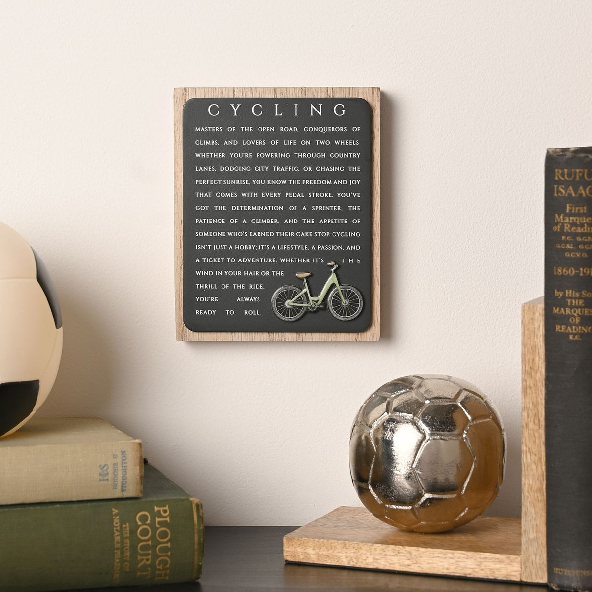Cycling Wooden Plaque with Green Bicycle in black and natural wood, displayed on a shelf beside books and a football, with white lettering text beneath the title.