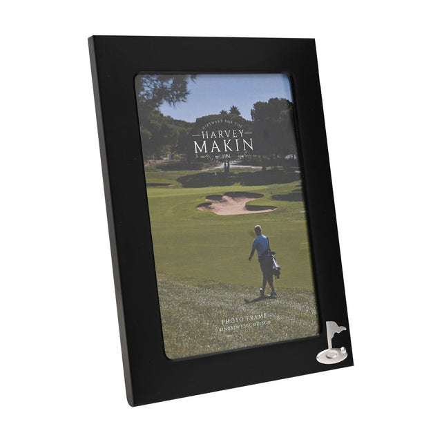 Rectangular matt black metal photo frame with a silver golf flag and ball emblem in the lower right corner, holding a 6 x 4in golf course photo, shown on a white background.