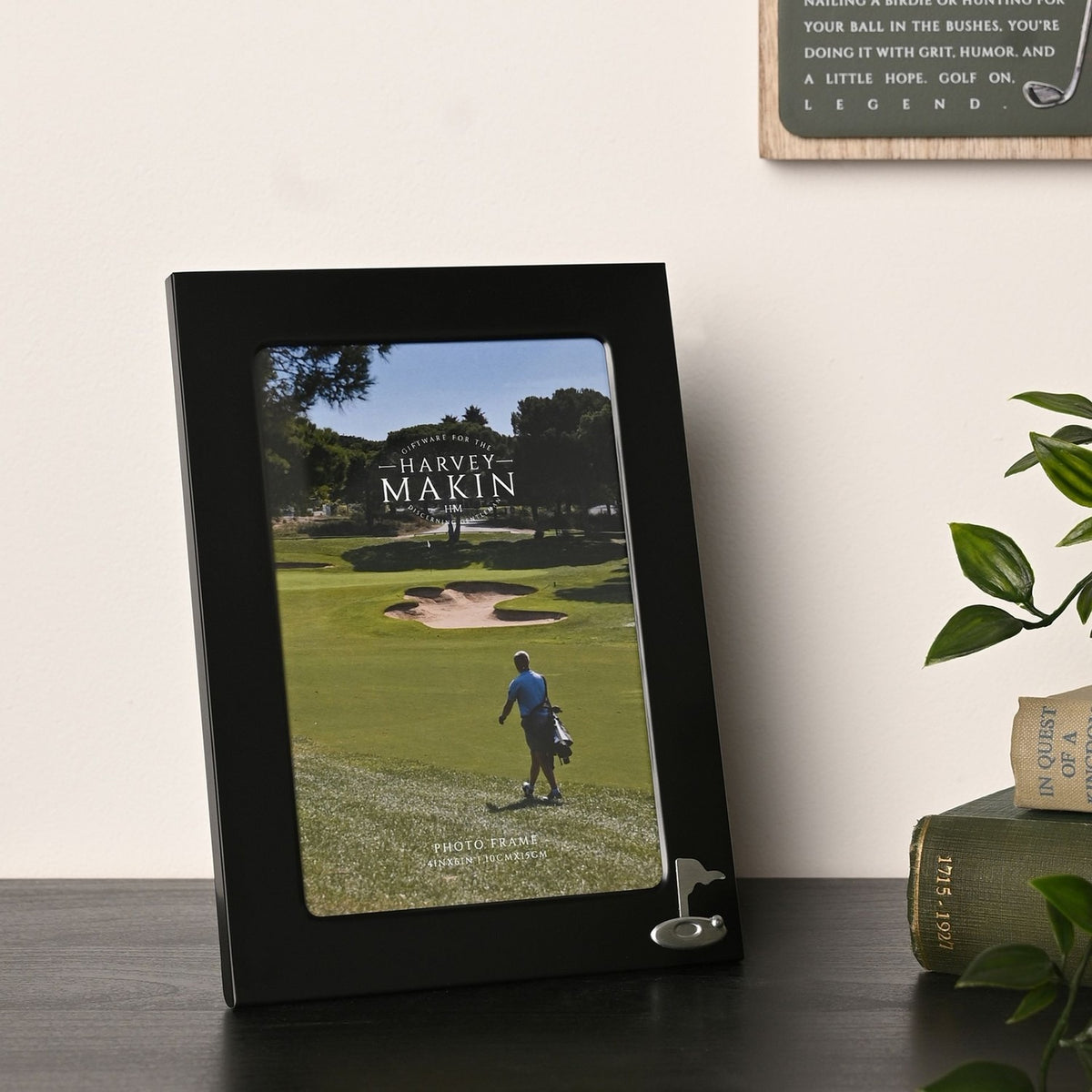 Matt black metal 6 x 4in photo frame with a silver golf flag emblem in the lower corner, shown on a dark shelf beside books and a plant, displaying a golf course photograph.
