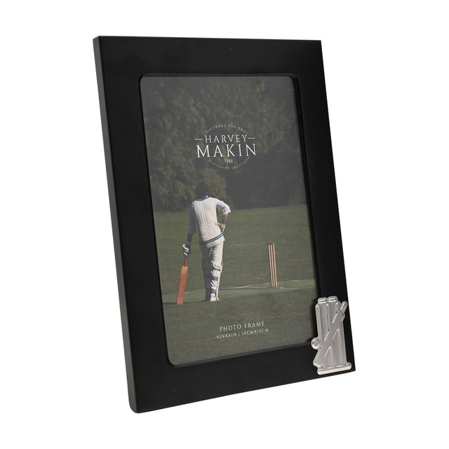 Black metal rectangular photo frame for a 6 x 4in picture, with a silver cricket stumps, bails and bat emblem on the lower right corner, shown on a white background.