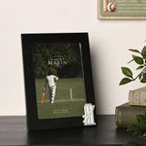 Black metal 6 x 4in photo frame with a silver cricket stumps, bails, bat and ball emblem on the lower right corner, shown on a dark shelf beside books and a plant.