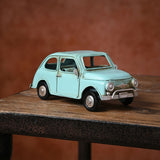 Classic car ornament, a pale aqua blue metal figurine with rounded bodywork, round headlamps and winged nose badge, shown on a wooden table against a rust-coloured background.