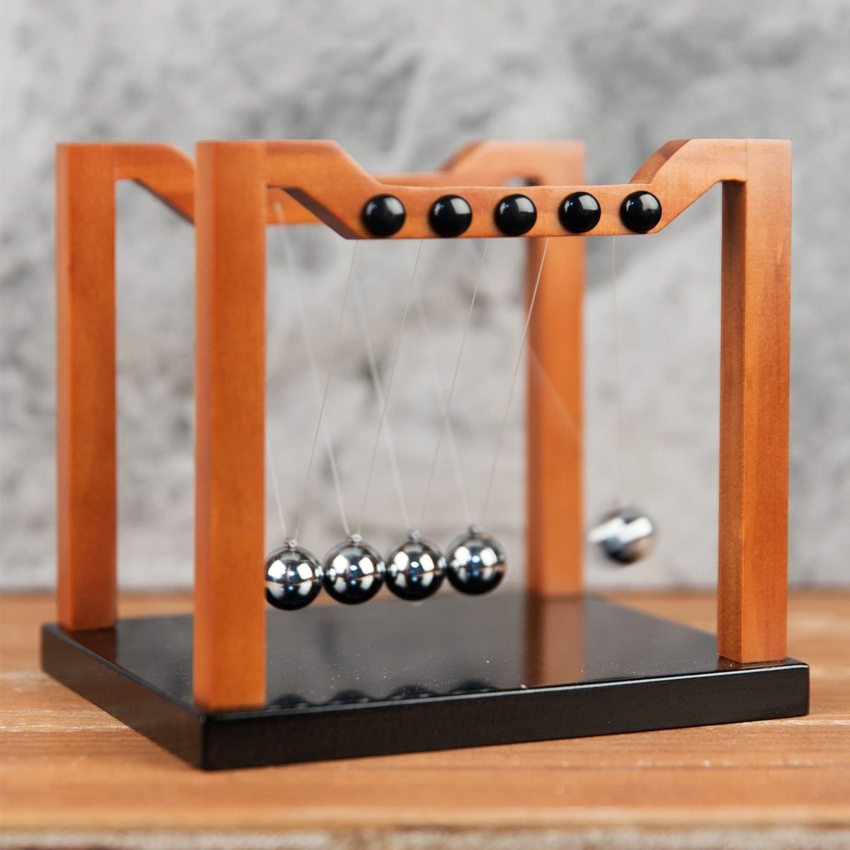 Wooden Newton's Cradle with black base, brown matt-finish frame, and five silver-coloured balls suspended on clear threads, shown on a wooden surface against a grey background.