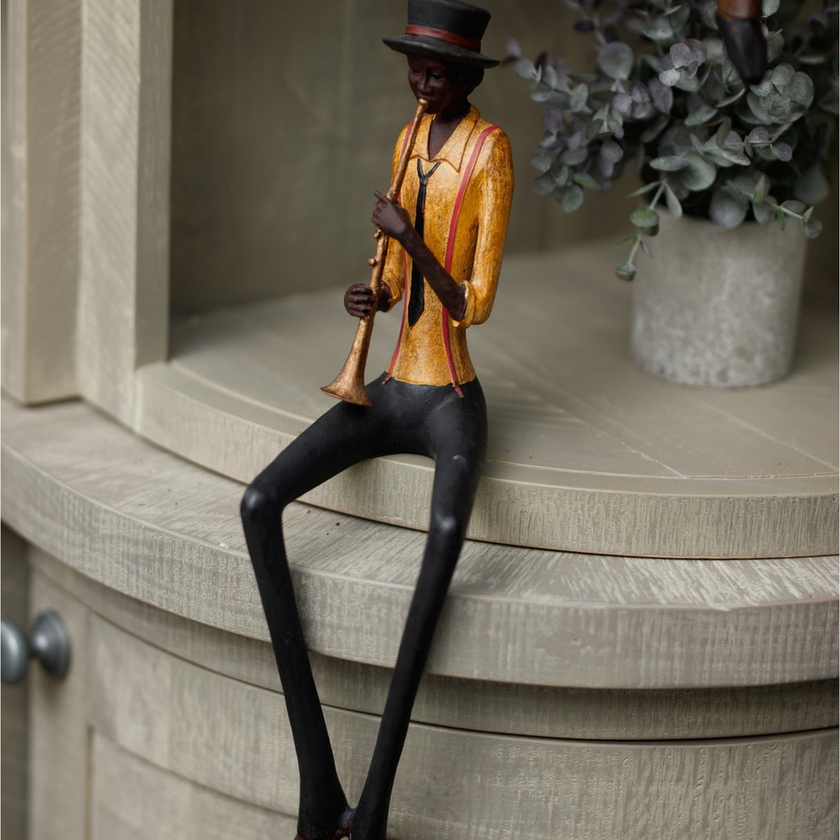 Hill Interiors Sitting Trumpeter Figurine in brown, seated resin musician in top hat and gold jacket playing trumpet, displayed on a shelf beside eucalyptus.
