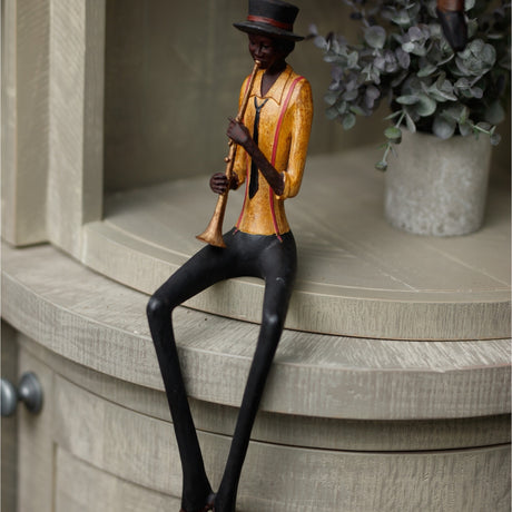 Hill Interiors Sitting Trumpeter Figurine in brown, seated resin musician in top hat and gold jacket playing trumpet, displayed on a shelf beside eucalyptus.