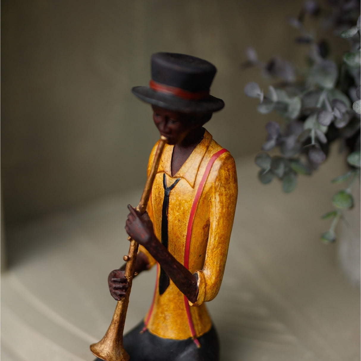 Hill Interiors Sitting Trumpeter Figurine in brown resin, shown playing a trumpet in a yellow jacket and black top hat beside eucalyptus foliage.