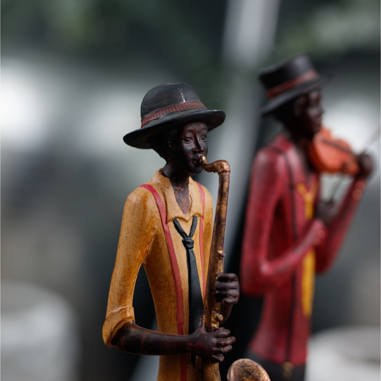 Hill Interiors Standing Saxophonist Figurine in brown resin, wearing a hat and yellow jacket, with a violinist figurine blurred behind.