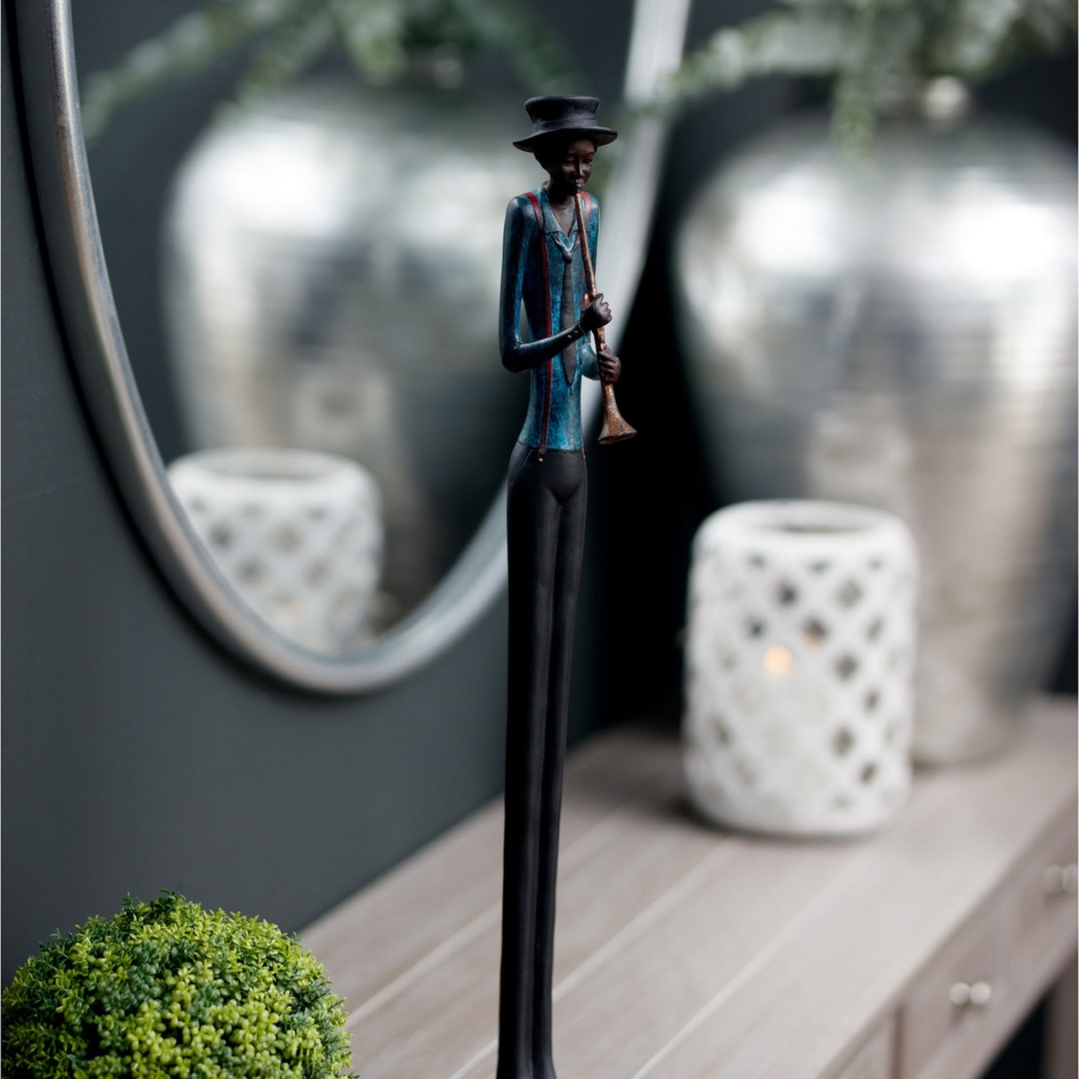 Hill Interiors Standing Trumpeter Figurine in resin with black hat, blue jacket and slim brown legs, displayed on a console beside a round mirror.