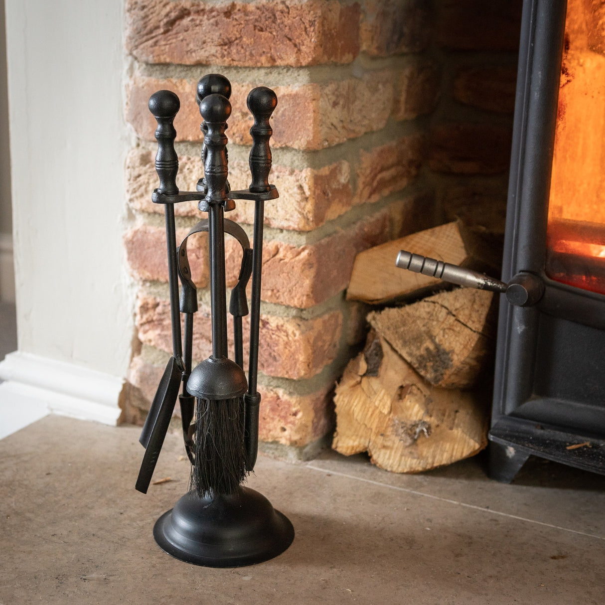 Hill Interiors Black Fireside Companion Set with poker, brush, shovel and tongs on an upright stand, placed beside a log-filled brick hearth and stove.