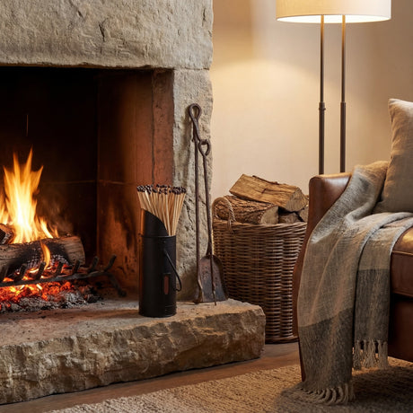 Hill Interiors black matchstick holder filled with 60 long matches, set on a stone hearth beside a lit fire, iron fire tools and wicker log basket.