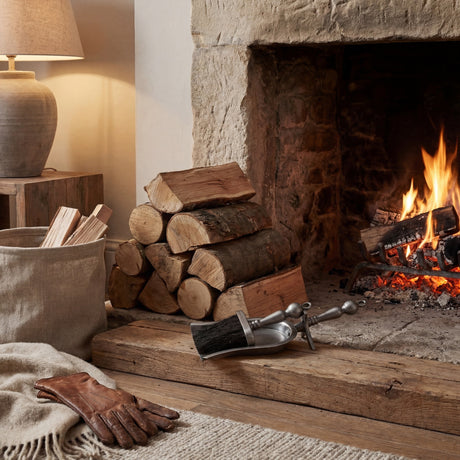 Hill Interiors antique pewter effect hearth brush and shovel resting on a wooden hearth beside stacked logs and a lit stone fireplace.