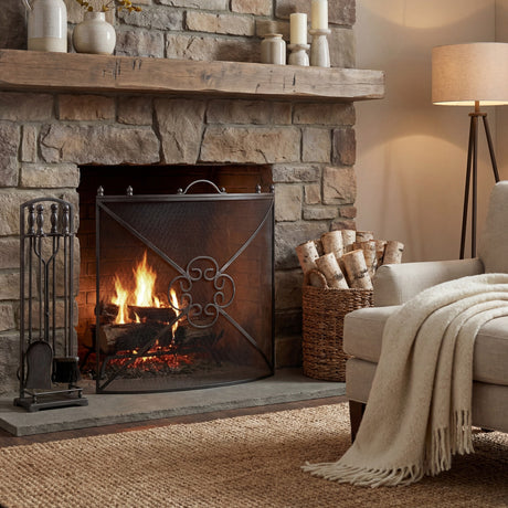 Hill Interiors mesh fireguard in antique pewter effect finish with scrolled centre motif, set before a lit stone fireplace in a cosy living room.