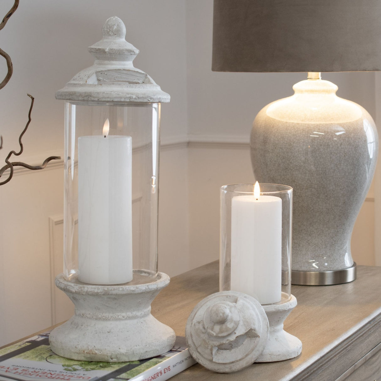 Hill Interiors Glass and Stone Pillar Candle Holder in white styled on a console with a lit pillar candle beside a smaller glass hurricane and lamp.