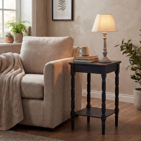 Hill Interiors wooden table lamp with turned base and beige tapered shade, styled on a black side table beside a cream armchair.