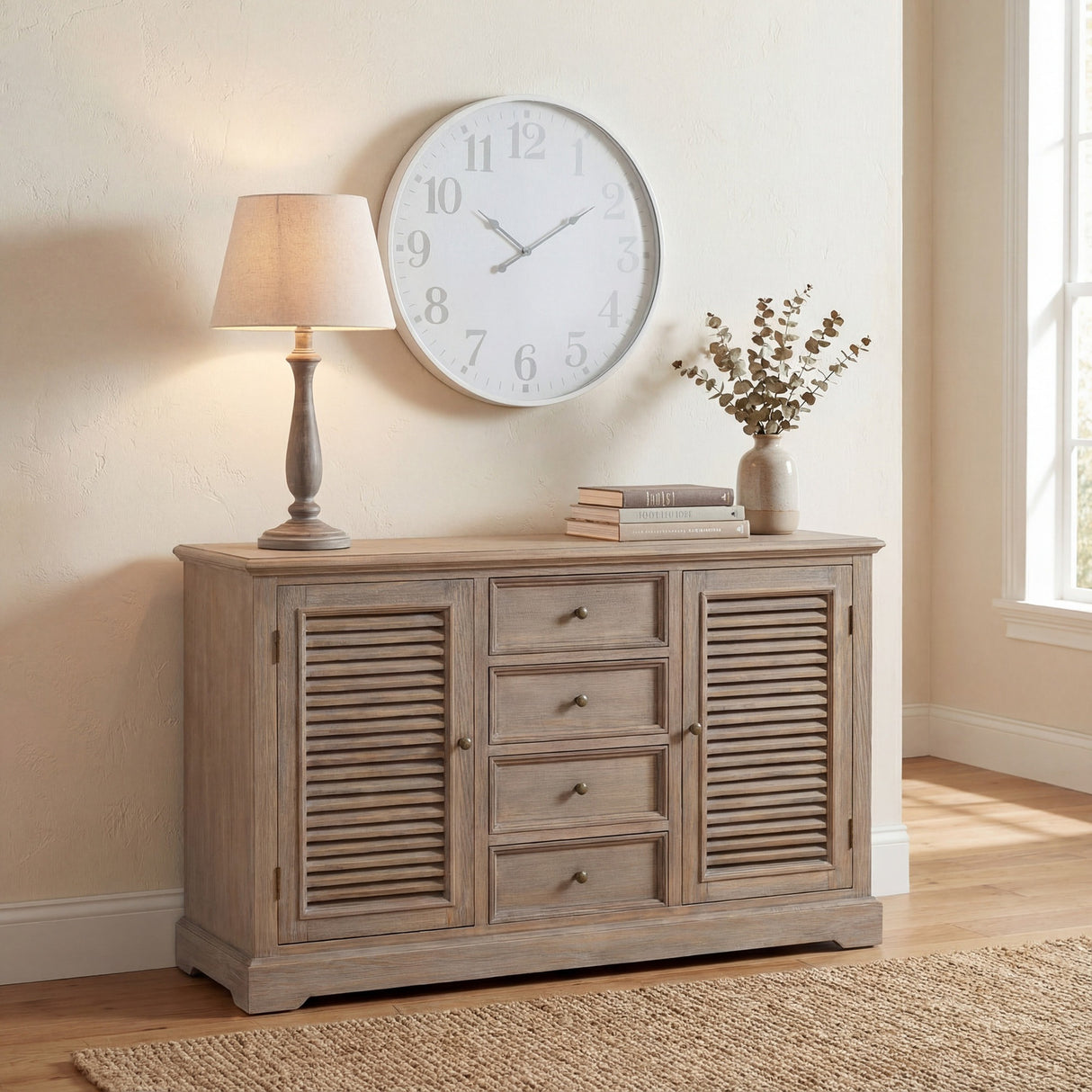 Hill Interiors Wooden Table Lamp in beige with a turned base and tapered fabric shade, lit on a wooden sideboard beside a wall clock and dried foliage.