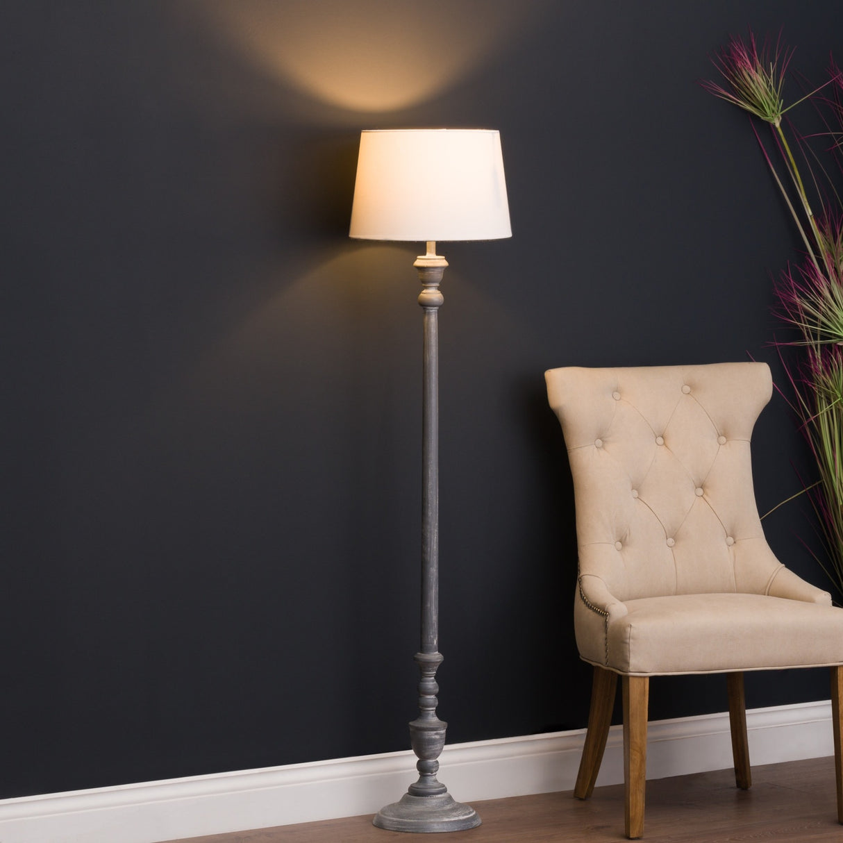 Hill Interiors wooden floor lamp with turned beige column and cream drum shade, lit beside a tufted beige chair against a dark wall.