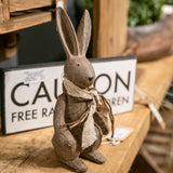 Hill Interiors Small Winter Bunny Rabbit Figurine in brown resin, upright with tall ears and a linen scarf, displayed on wood beside a Caution sign.