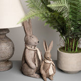Hill Interiors Small Winter Bunny Rabbit Figurine in brown resin, shown with a taller bunny beside a lamp base and potted fern on a console.