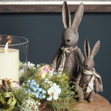 Hill Interiors Large Winter Bunny Rabbit Figurine in brown resin, shown with a smaller bunny beside a candle and floral arrangement on a wooden table.