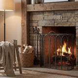 Hill Interiors Gothic fire screen in antique pewter finish, arched tracery panels set before a lit stone fireplace with log basket and companion set.