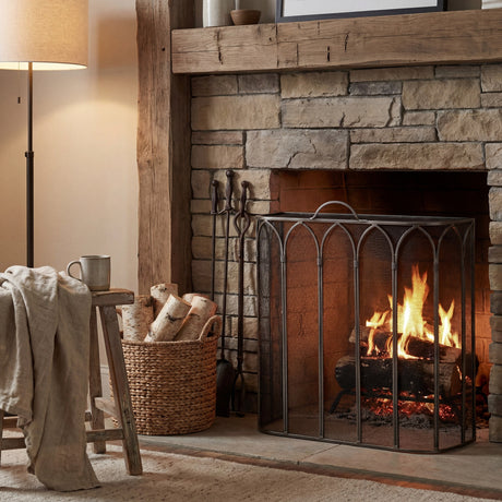Hill Interiors Gothic fire screen in antique pewter finish, arched tracery panels set before a lit stone fireplace with log basket and companion set.