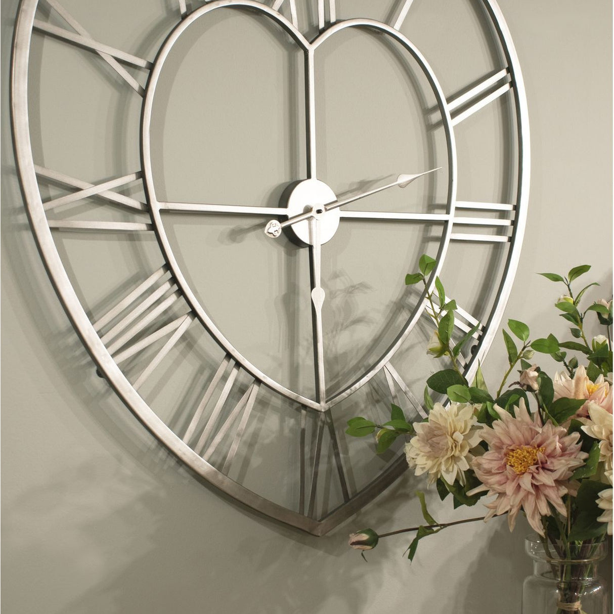 Hill Interiors Skeleton Heart Wall Clock in silver metal with openwork Roman numerals, shown beside a vase of pink and cream flowers.