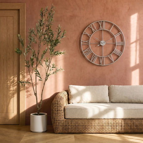Hill Interiors Large Silver Skeleton Wall Clock with Roman numerals mounted on a terracotta wall above a rattan sofa beside a potted olive tree.