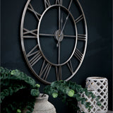 Hill Interiors large skeleton wall clock with silver foil finish, open Roman numerals and slim hands, mounted on a dark wall above greenery and a lantern.