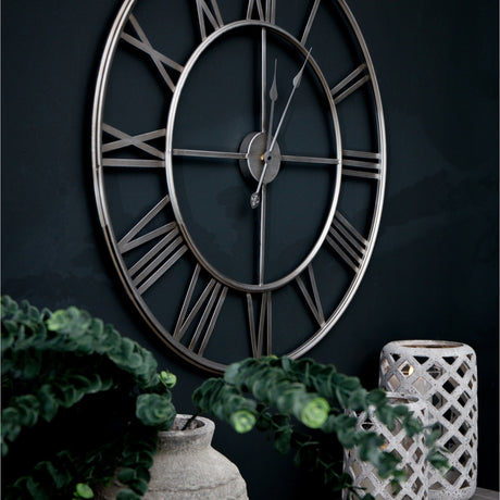 Hill Interiors large skeleton wall clock with silver foil finish, open Roman numerals and slim hands, mounted on a dark wall above greenery and a lantern.