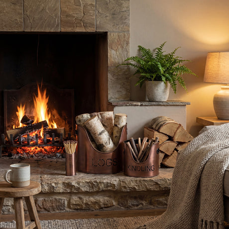 Hill Interiors copper finish log and kindling buckets with matchstick holder set on a stone hearth beside a lit fireplace and stacked logs.
