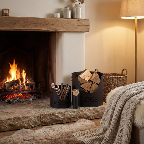 Hill Interiors black metal log and kindling buckets with matchstick holder set on a stone hearth beside a lit fireplace and woven basket.