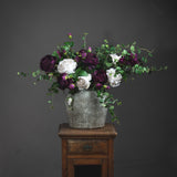 Hill Interiors Aged Stone Ceramic Vase in beige with rounded body holding purple and white roses with trailing eucalyptus, on a wooden stand.