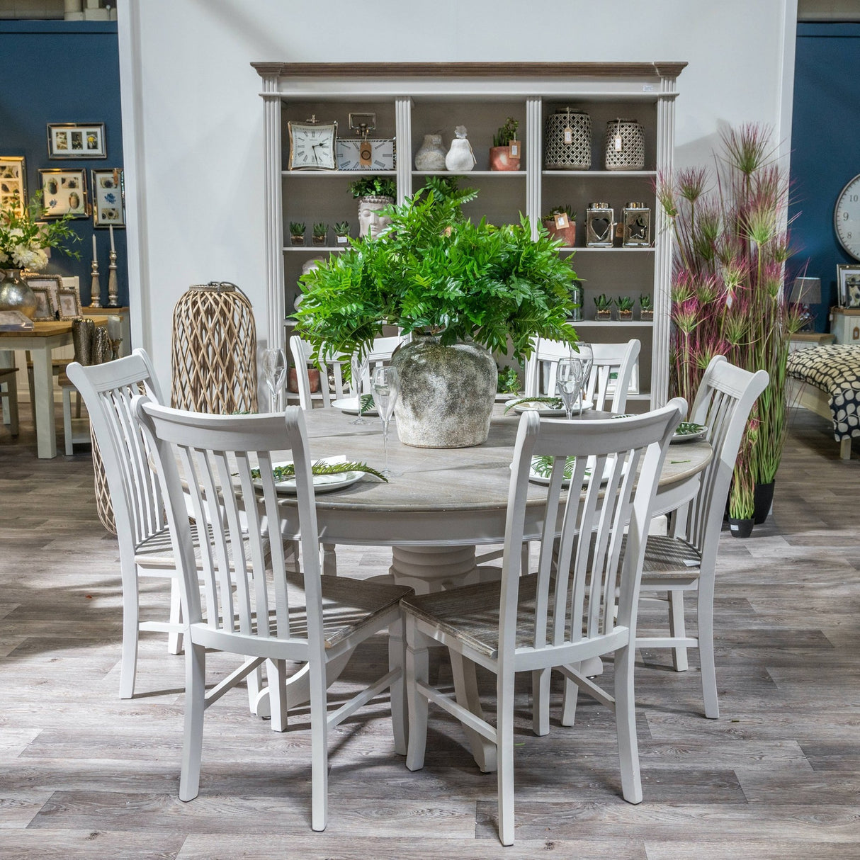 Hill Interiors Aged Stone Ceramic Vase in beige with lush green foliage, centred on a round dining table set with glassware and white chairs.
