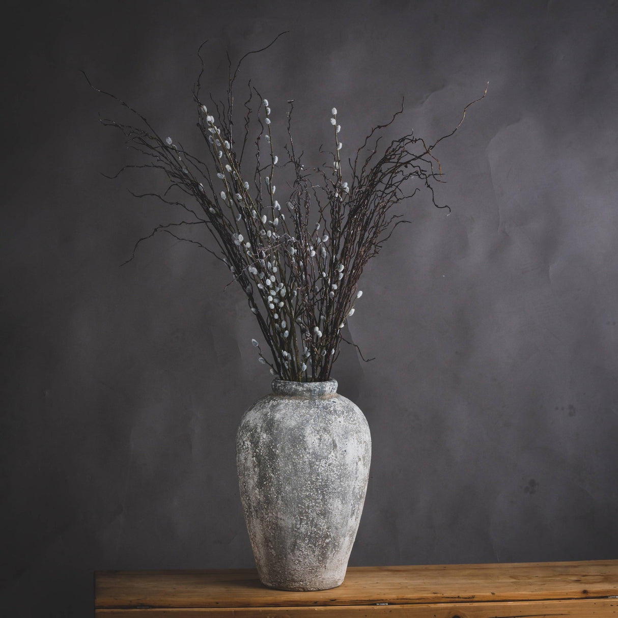 Hill Interiors tall beige ceramic vase with aged stone finish, holding pussy willow branches, displayed on a wooden surface against a dark grey backdrop.