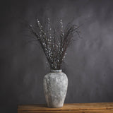 Hill Interiors tall beige ceramic vase with aged stone finish, holding pussy willow branches, displayed on a wooden surface against a dark grey backdrop.
