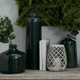 Hill Interiors large conical ceramic lattice hurricane lantern in beige, holding a lit candle, styled with dark green vases, books and ferns on a wooden shelf.