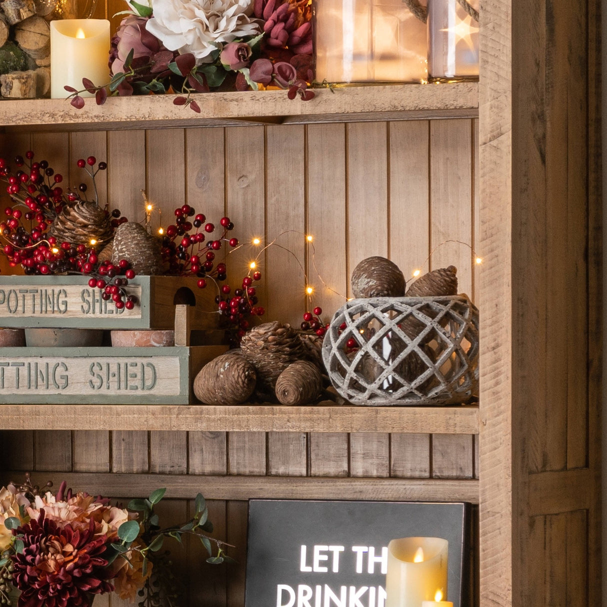 Hill Interiors round beige ceramic lattice hurricane lantern displayed on a rustic wooden shelf with pine cones, red berries and fairy lights.