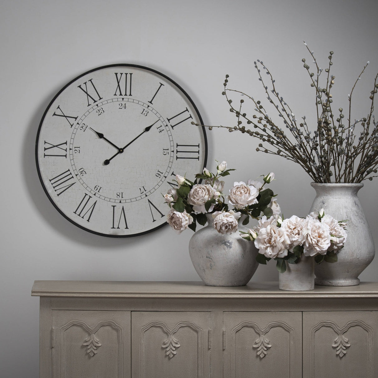 Hill Interiors Large Station Wall Clock in white with black Roman numerals and slim hands, mounted above a grey sideboard styled with stone vases and roses.