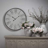 Hill Interiors Large Station Wall Clock in white with black Roman numerals and slim hands, mounted above a grey sideboard styled with stone vases and roses.