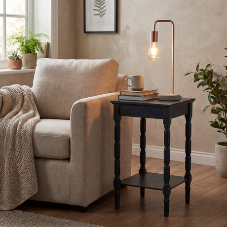 Hill Interiors copper industrial table lamp with exposed bulb and stone base on a black turned-leg side table beside a beige armchair in a living room.