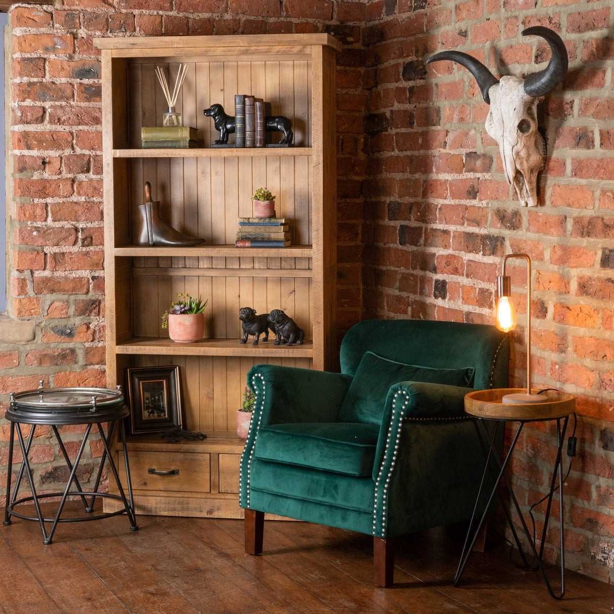Hill Interiors copper industrial table lamp with exposed bulb on a round wooden tray table beside a green velvet armchair against a brick wall.